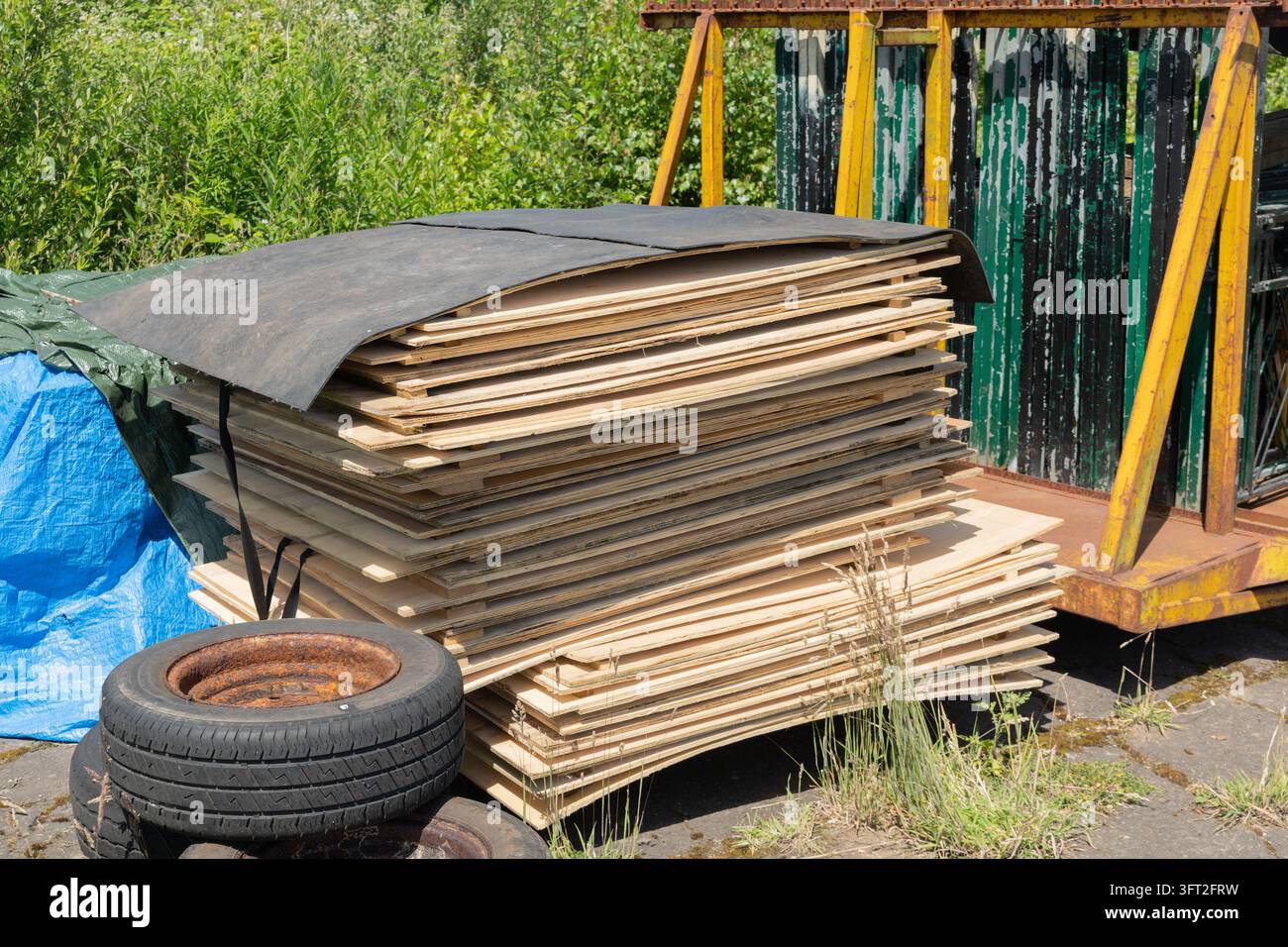 Großer Stapel flacher Holztafeln, bedeckt mit schwarzer Plane, gelagert neben alten Reifen und blauer Plastikfolie auf einem Außenhof einer Farm setti Stockfoto