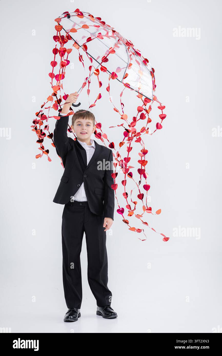Junge, formell gekleidet, hält einen Regenschirm mit glänzenden roten Herzen, lächelt und posiert mit weißem Hintergrund für ein Feier-Motto. Stockfoto