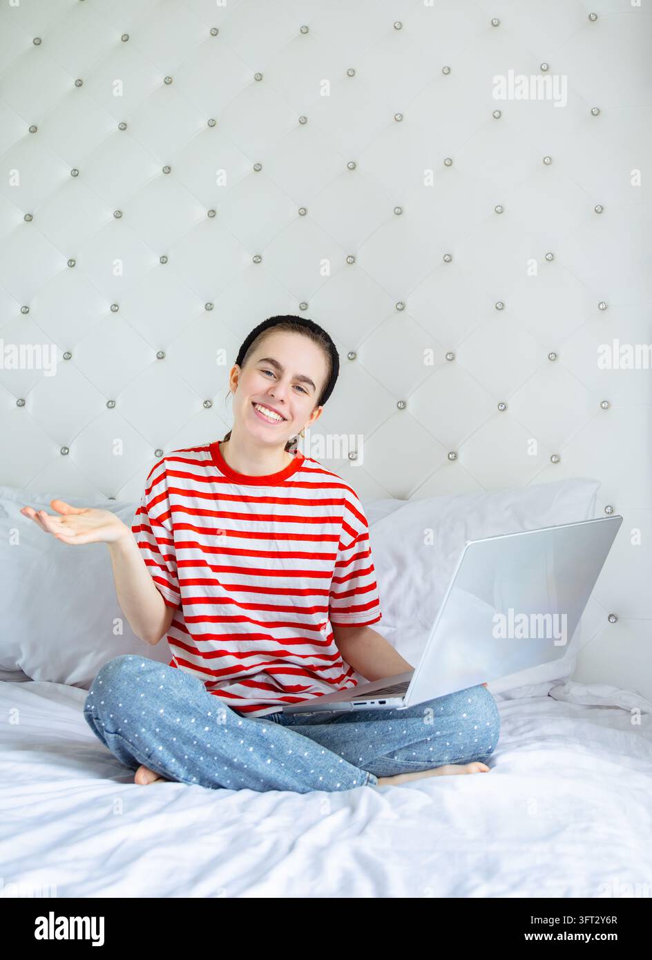 Lächelnde Frau, die einen Laptop benutzt, sitzt mit gekreuzten Beinen in entspannter, lässiger Kleidung auf einem weißen Bett in einer komfortablen Umgebung Stockfoto