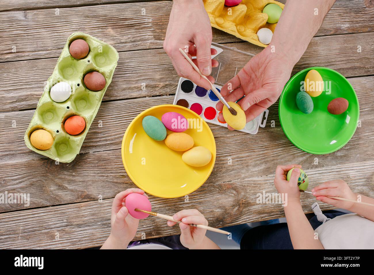 Helle Holzeier, die mit verschiedenen Farben bemalt sind, werden von Händen mit Pinseln gehalten, bunte Teller und Malpalette sind sichtbar. Stockfoto