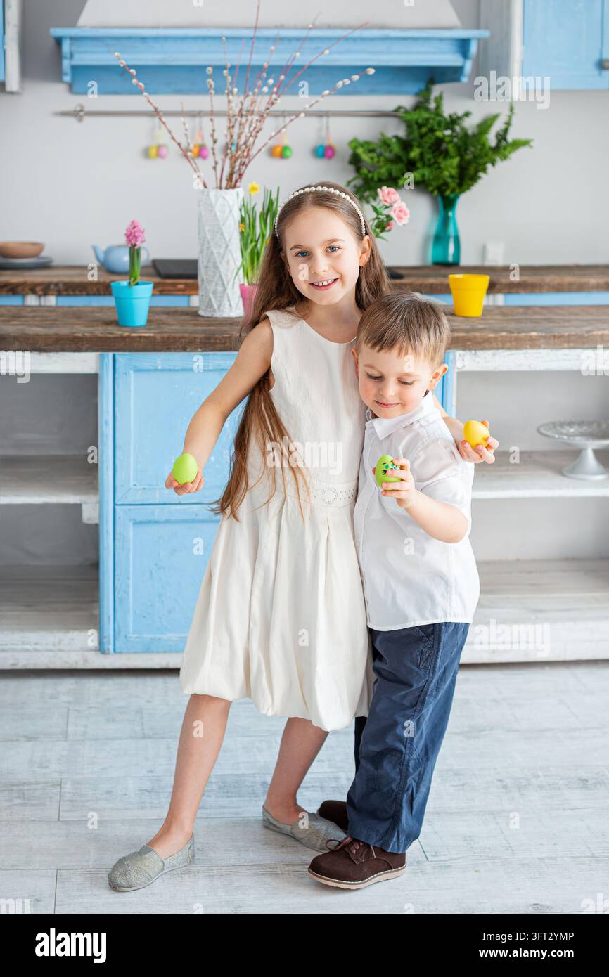 Lächelnde Kinder halten bunte Ostereier, umgeben von Frühlingsdekorationen und hellem Raum. Stockfoto