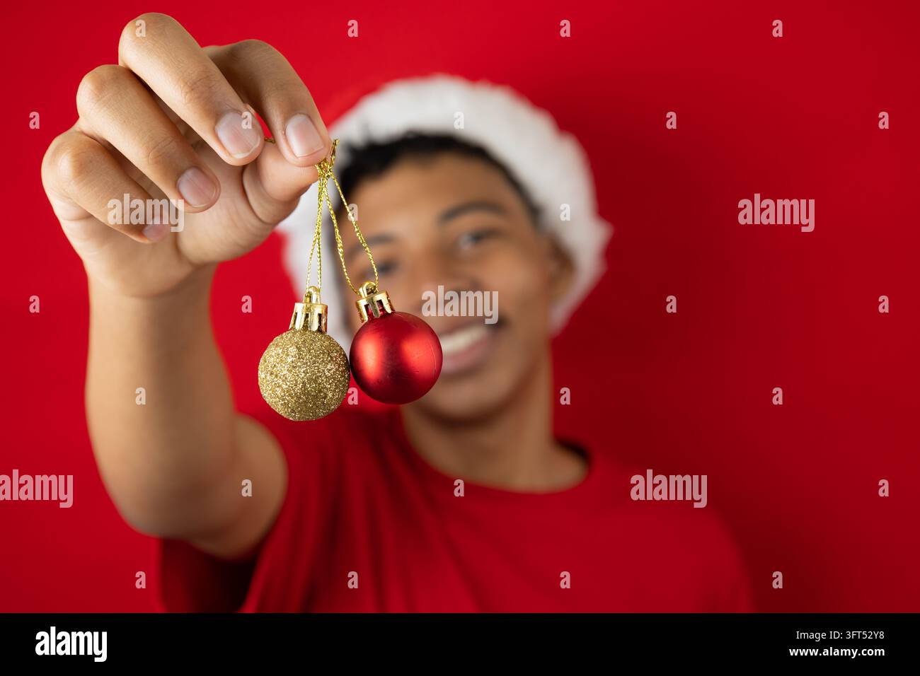 Unscharfer dunkelhäutiger Teenager hält zwei Weihnachtsschmuckstücke, eines rot und eines gold, vor einem roten Hintergrund. Fröhliches festliches Bild, ideal für Design Stockfoto