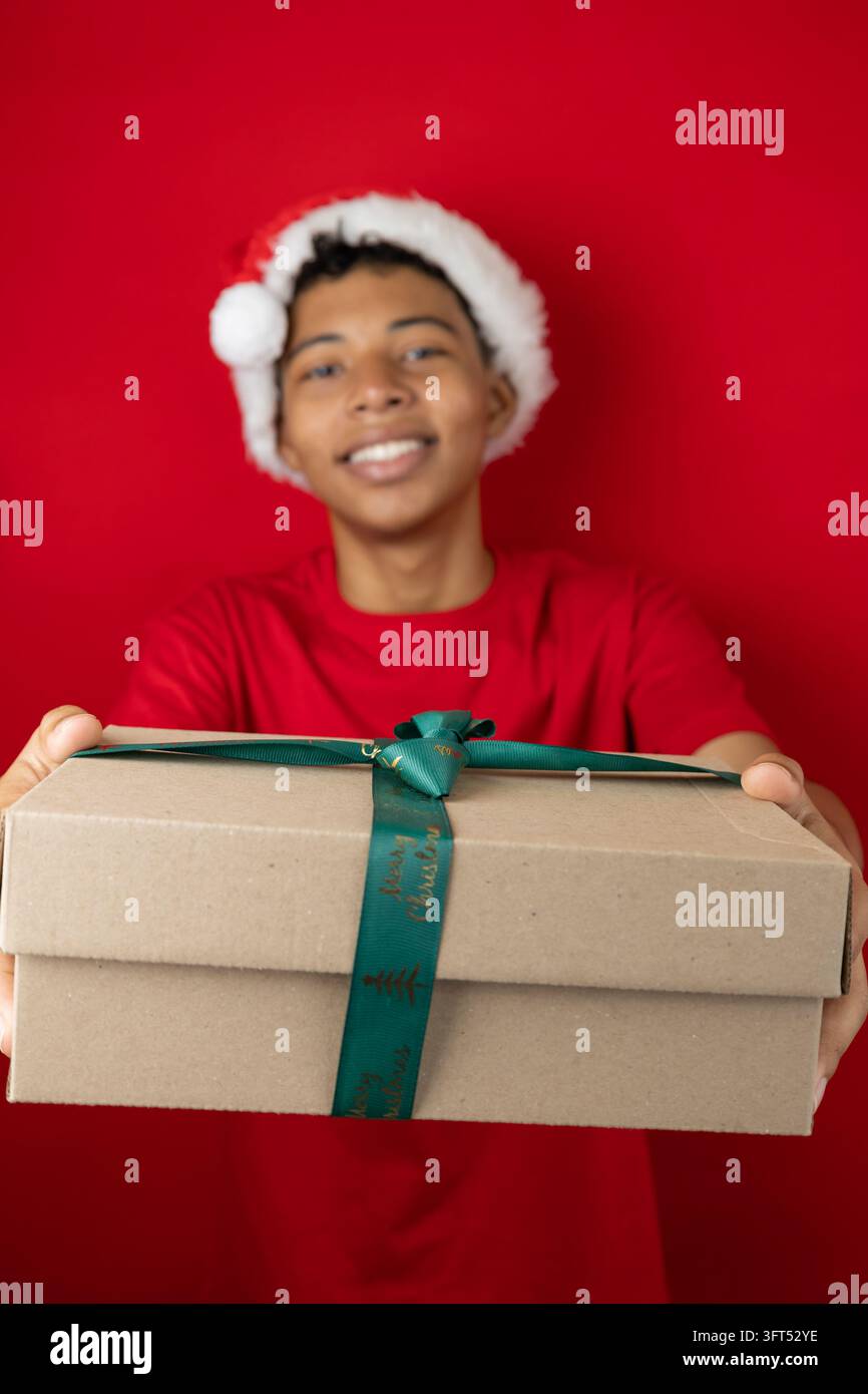 Unscharfer dunkler junger Mensch, der eine Geschenkschachtel hält und anbietet, vor rotem Hintergrund mit Platz für Text. Festliches und modernes Bild, ideal für de Stockfoto