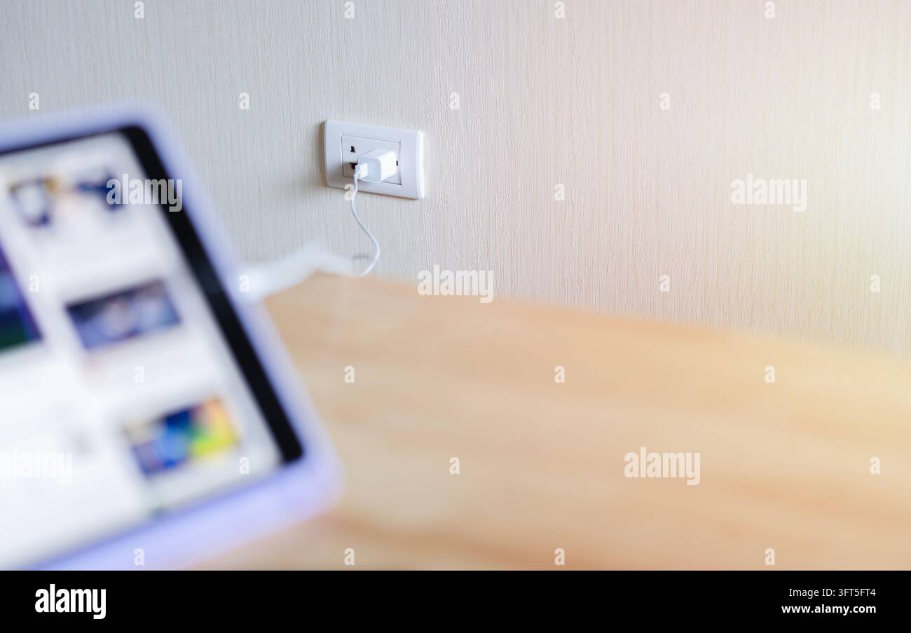 LadeDigitales Tablet mit einem Kabel auf hellem beigem Hintergrund. Telefon an Ladegerät angeschlossen. Stockfoto