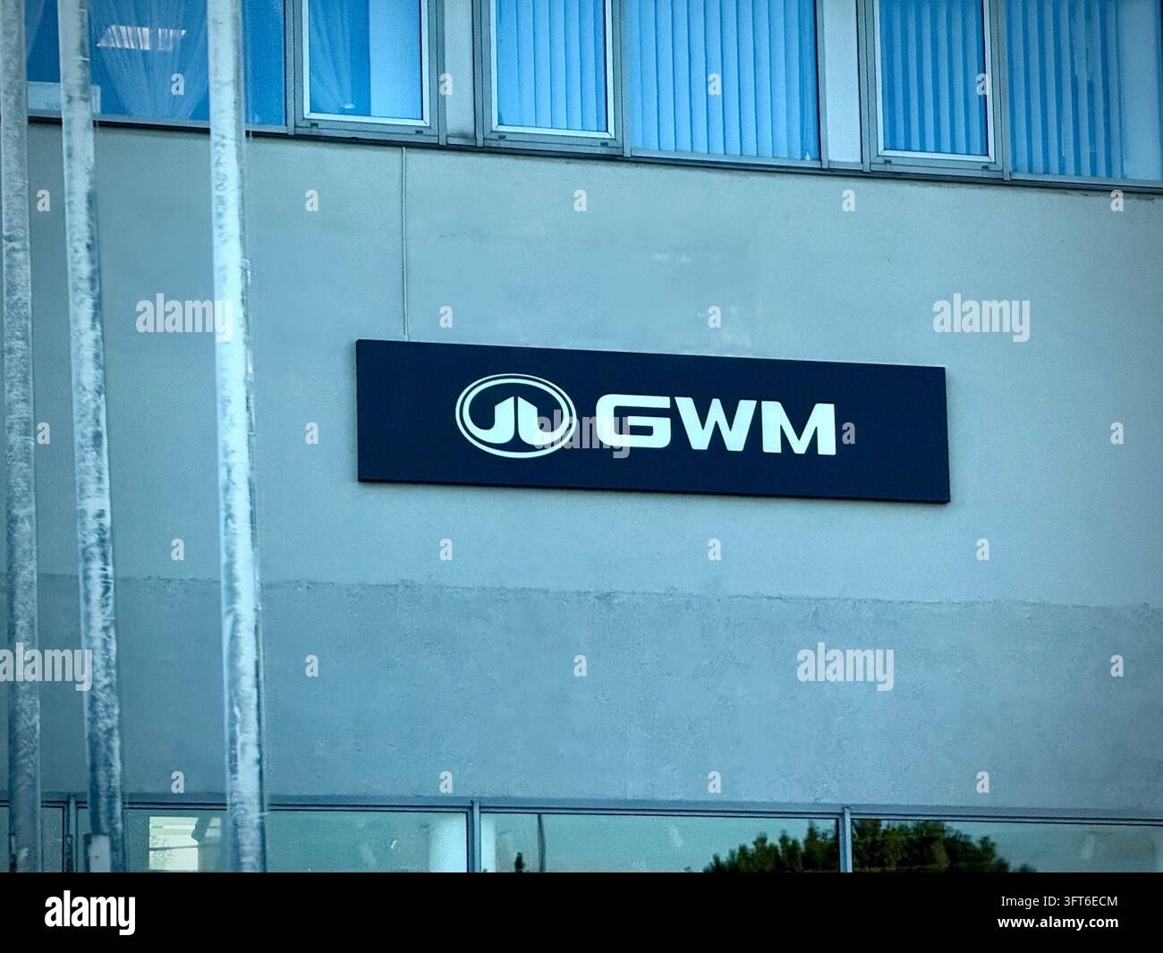 weißes GWM-Markenzeichen und Firmenlogo, das auf einer hellgrauen Gebäudefassade über Glasfenstern montiert ist, mit vertikalen Stangen links. - Smartphone-aufgenommenes Stockfoto