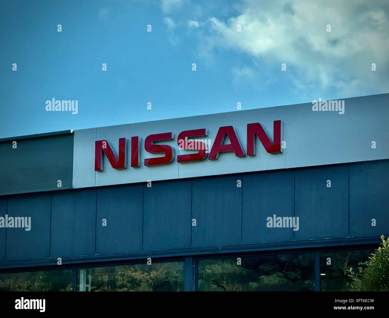 Rote Nissan-Markenschilder montiert auf einem hellgrauen Rahmen über einem modernen Autohandelssaal, vor einem hellblauen Himmel mit weichen weißen Wolken. - Smartphone-aufgenommenes Stockfoto
