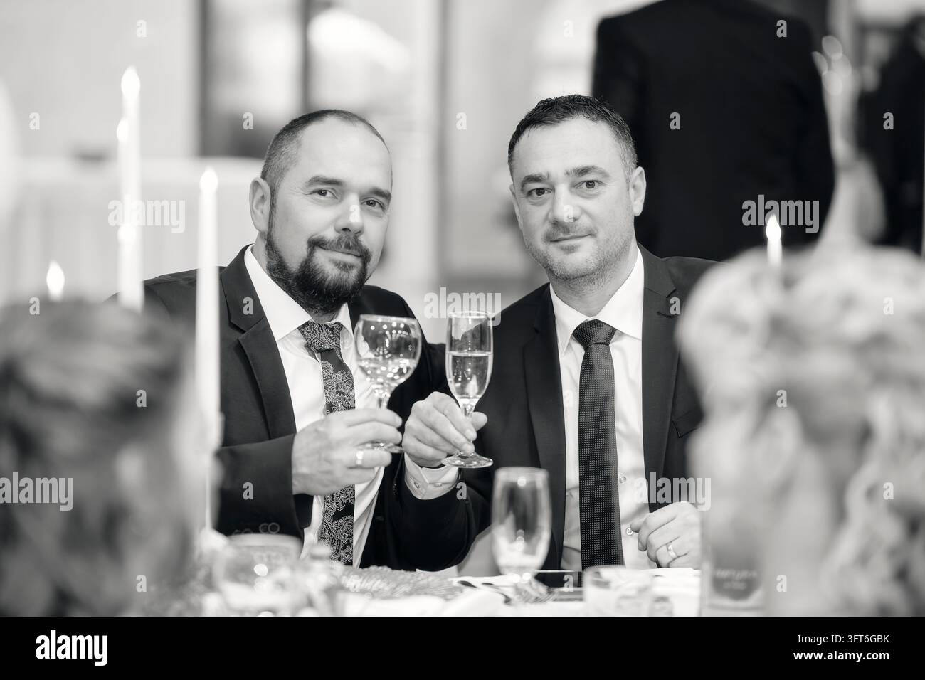 Schwarz-Weiß-Porträt von zwei Männern in Anzügen, die Stielgläser heben, während sie an einem kerzenbeleuchteten Balltisch sitzen. Stockfoto