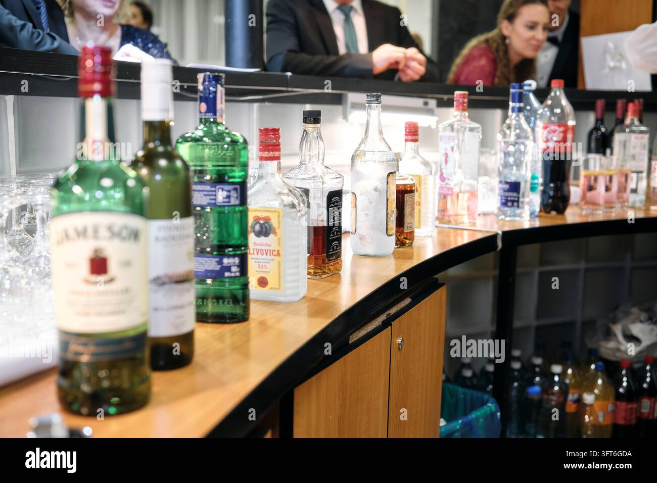 Flaschen mit Spirituosen und alkoholfreien Getränken säumen eine gebogene Holztheke, wobei Gäste in einem Spiegel während eines Balls gespiegelt werden. Stockfoto