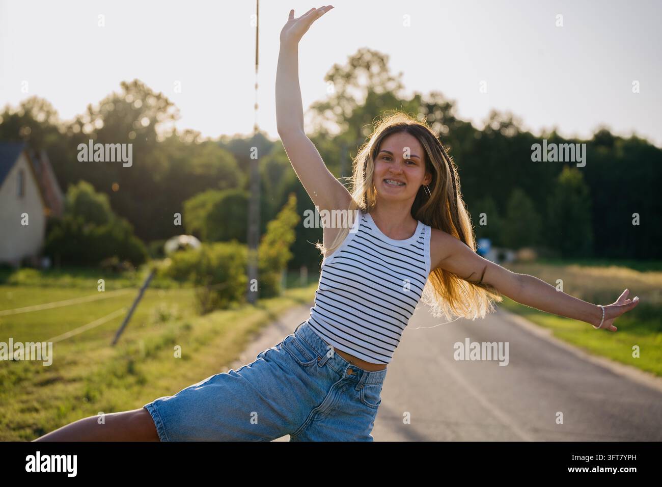 Lächelnde junge Frau tanzt mit erhobenen Armen auf einer ruhigen Landstraße bei Sonnenuntergang. Emotionales Wohlbefinden, verspielte Erwachsenenzeit und freudige Freiheit. Stockfoto
