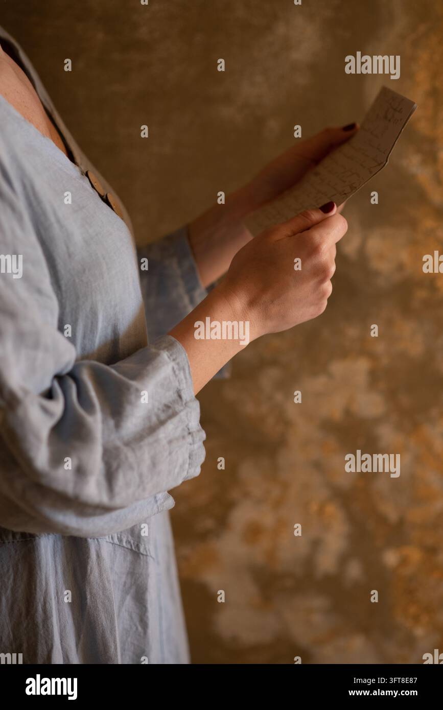 Eine Frau hält einen kleinen handgeschriebenen Zettel in beiden Händen, mit rotem Nagellack und einem hellgrauen Bluse vor einem warmen Steinhintergrund. Stockfoto