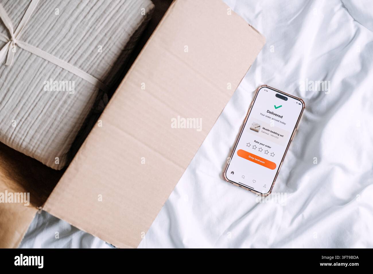 Telefon, das die Ankunft einer Bettbestellung neben einem offenen Paket mit gefaltetem Baumwollgaze bestätigt, veranschaulicht die schnelle Lieferung nach Hause, die über die mobile App verfolgt wird Stockfoto