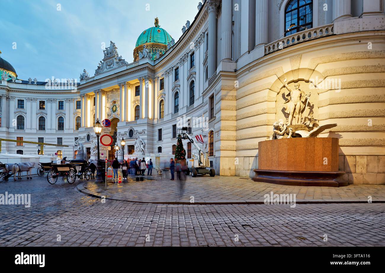 Fiaker-Kutschen und Fußgänger passieren den kaiserlichen Michaelertrakt und seinen Brunnen, während Anfang November ein Weihnachtsbaum aufgestellt wird, Österreich. Stockfoto