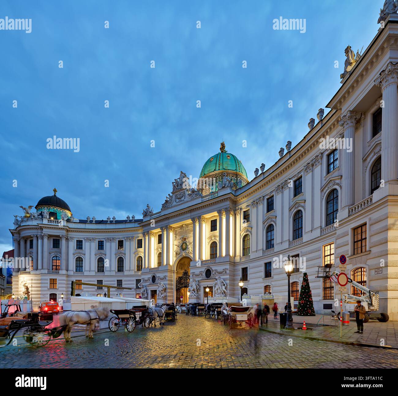 Fiaker Pferdekutschen warten auf dem gepflasterten Michaelerplatz vor dem beleuchteten St. Michaelstor und der grünen Kuppel, mit einem Weihnachtsbaum zur Blauen Stunde. Stockfoto