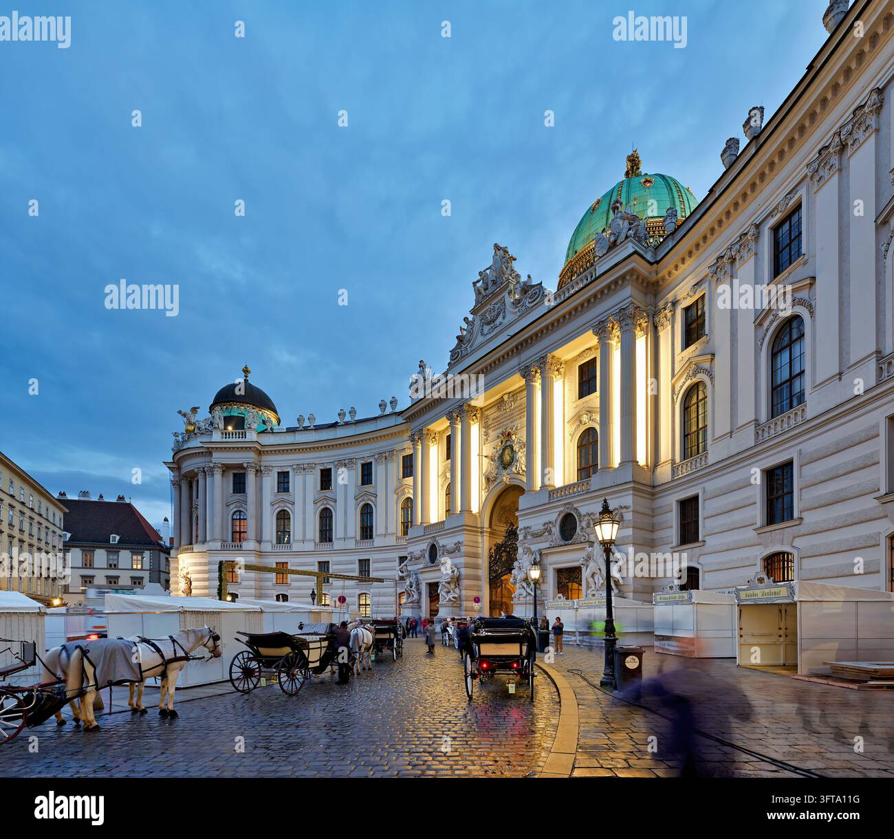 Pferdekutschen warten auf nassen Kopfsteinpflaster des Michaelerplatzes unterhalb des St. Michael-Flügels des kaiserlichen Palastes, Wien, Österreich, in der Blauen Stunde Stockfoto