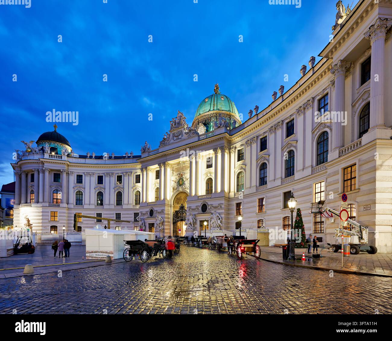 Fiaker-Kutschen warten auf nassem Kopfsteinpflaster vor dem beleuchteten St. Michaels-Flügel des Hofburg-Imperpalastes, Wien, Österreich, in der Blauen Stunde. Stockfoto