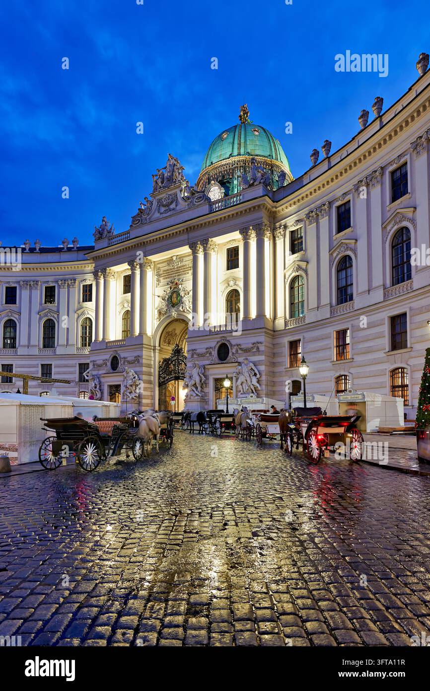 Pferdegespanne warten auf nassen Kopfsteinpflaster des Michaelerplatzes vor dem beleuchteten St. Michael-Flügel der Hofburg, Österreich, zur Blauen Stunde. Stockfoto