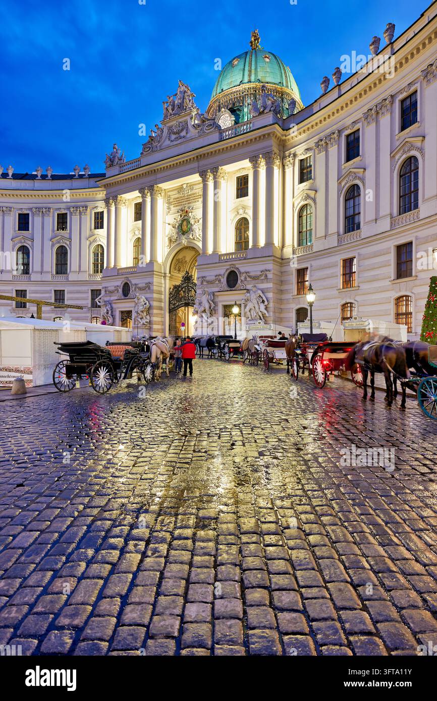 Pferdegespanne warten auf nassen Kopfsteinpflaster des Michaelerplatzes vor dem beleuchteten St. Michael-Flügel des kaiserlichen Palastes zur Blauen Stunde, Österreich Stockfoto
