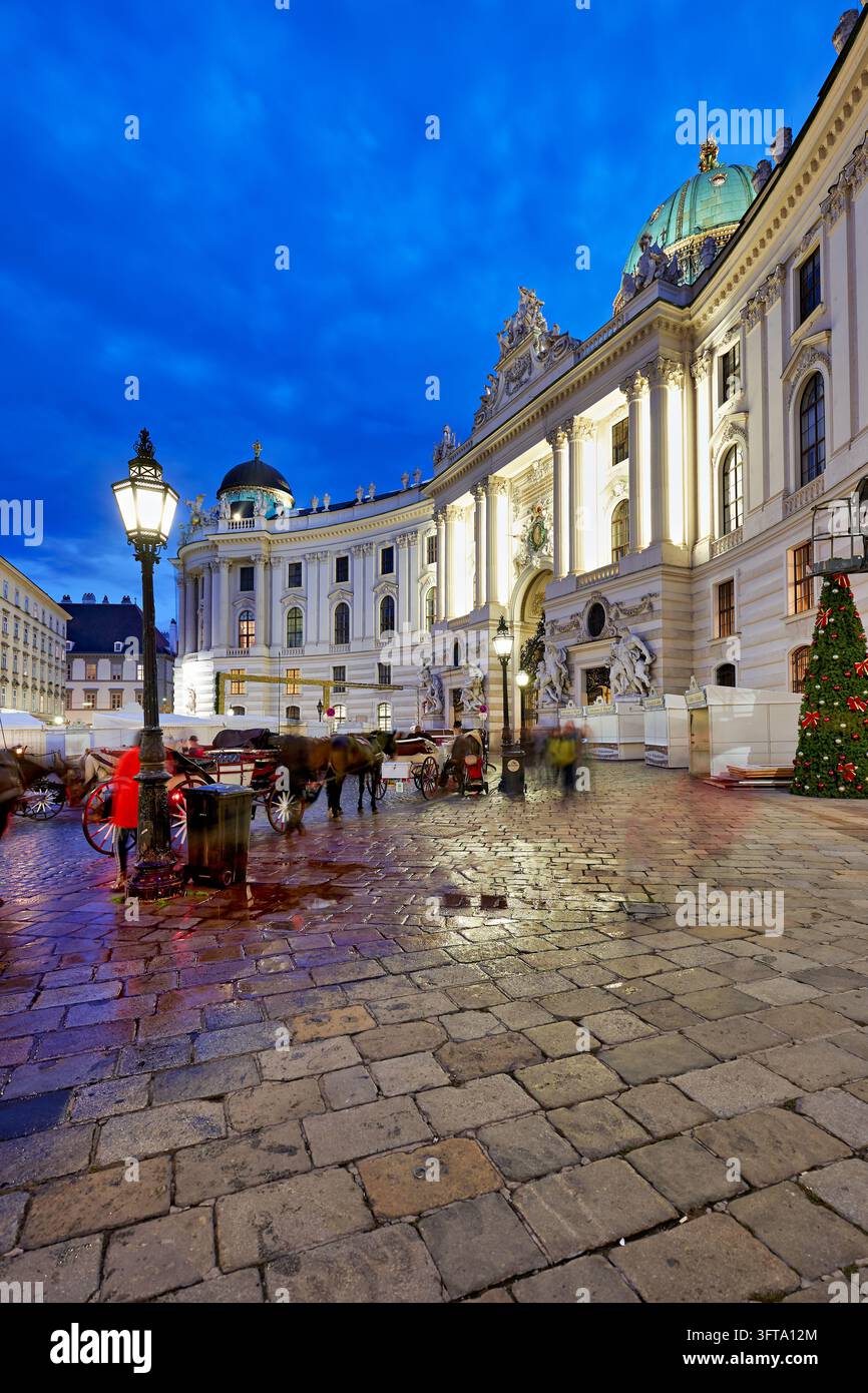Pferdekutschen warten auf nassen Kopfsteinpflaster des Michaelerplatzes in der Blauen Stunde neben einem Weihnachtsbaum und geschlossenen Marktständen, Wien, Österreich Stockfoto