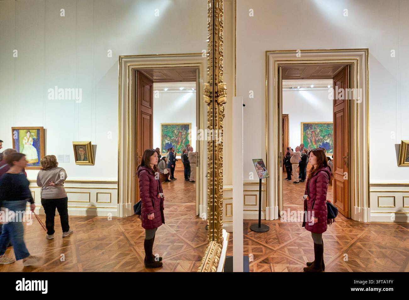 Eine Frau in einem kastanienbraunen Mantel steht vor einem vergoldeten Spiegel in einer Galeriehalle mit Parkettböden, während Besucher Gemälde betrachten, Wien, Österreich. Stockfoto