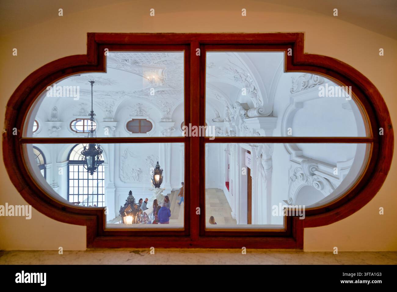Besucher steigen eine weiße, stuckverkleidete Große Treppe mit Laternen hinauf, gesehen durch einen ovalen Holzrahmen in einem historischen Gebäude in Wien, Österreich Stockfoto
