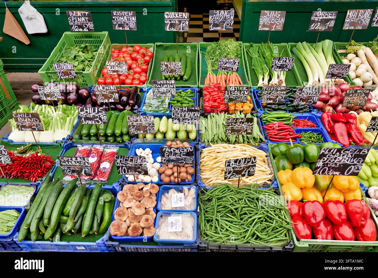 Frische Produkte wie Paprika, grüne Bohnen, Pilze und Gurken in Kisten mit handgeschriebenen deutschen Etiketten auf einem Lebensmittelmarkt in Wien. Stockfoto
