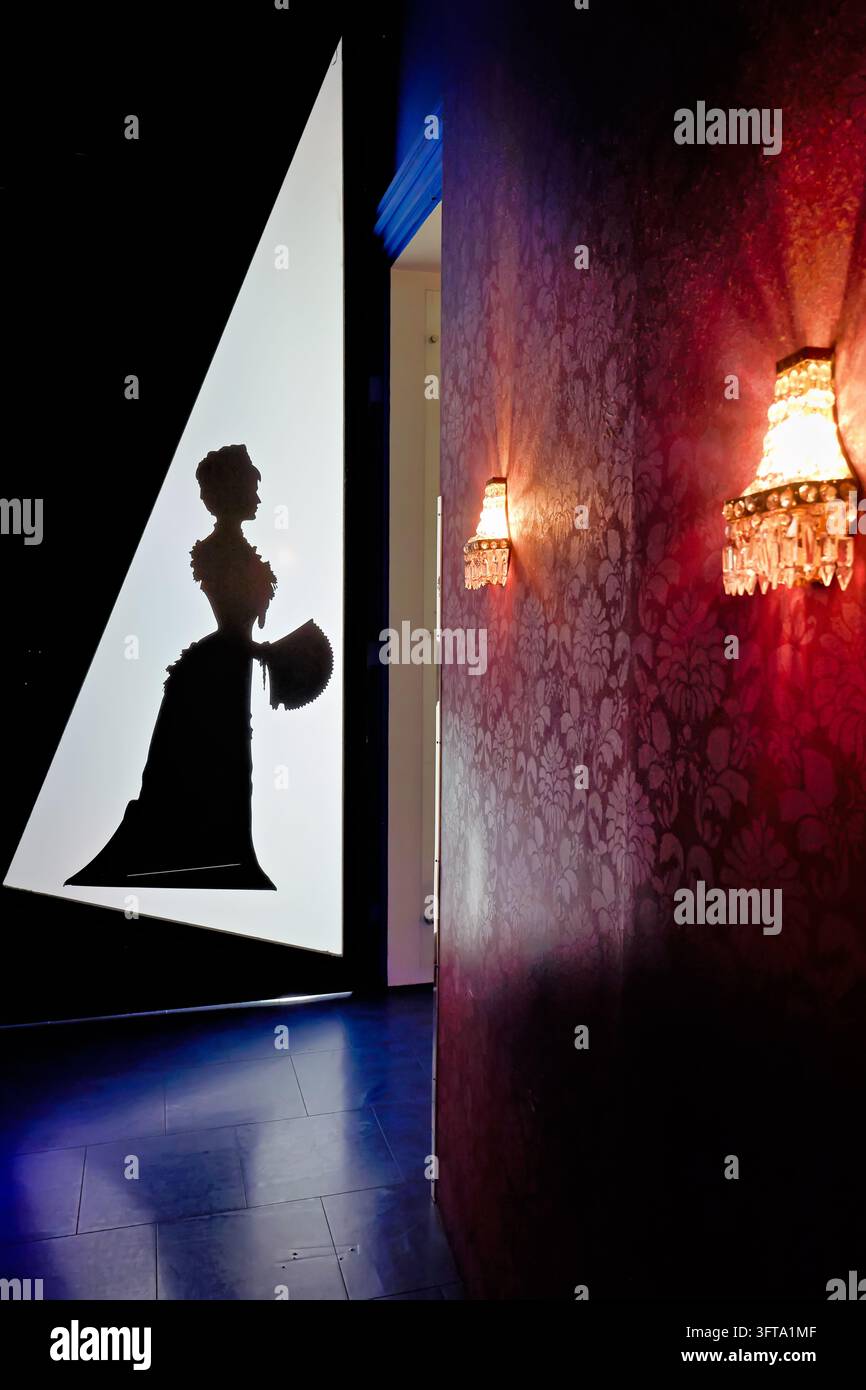Rückseitenbeleuchteter Ausschnitt einer Frau in einem Büstekleid, die einen Fächer neben Damasttapeten und Kristallwandleuchten in einem Ausstellungsraum in Wien, Österreich, hält Stockfoto