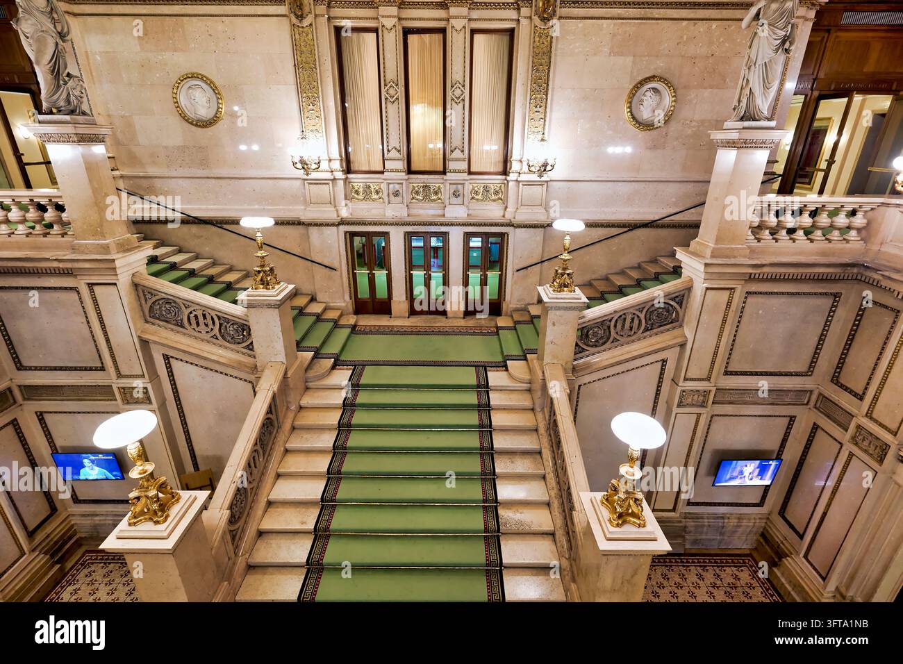 Symmetrische Ansicht von Marmorstufen mit grünen Teppichläufern, vergoldeten Lampen und Statuen im Wiener Staatsoper Opernhaus in Wien Stockfoto