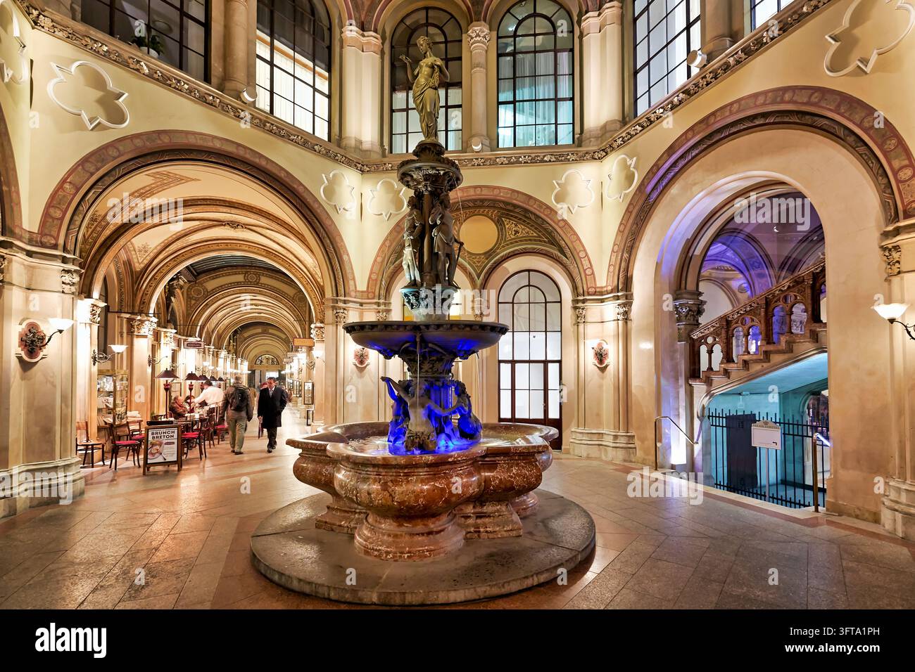 Der Donaudonauf-Brunnen leuchtet blau im Innenhof der historischen Einkaufspassage, während Fußgänger an einem Café an einem Herbstabend in Österreich vorbeigehen Stockfoto
