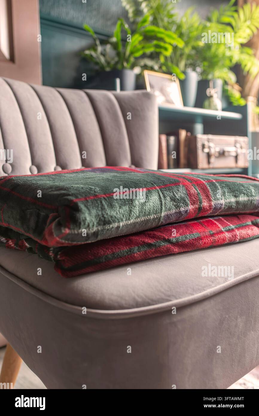 Grüne rote Karodecke aus Fleece, gefaltet auf einem grauen Stuhl in einem dunkelgrünen und goldenen Raum, mit Sonnenlicht, das in den Raum scheint. Stockfoto