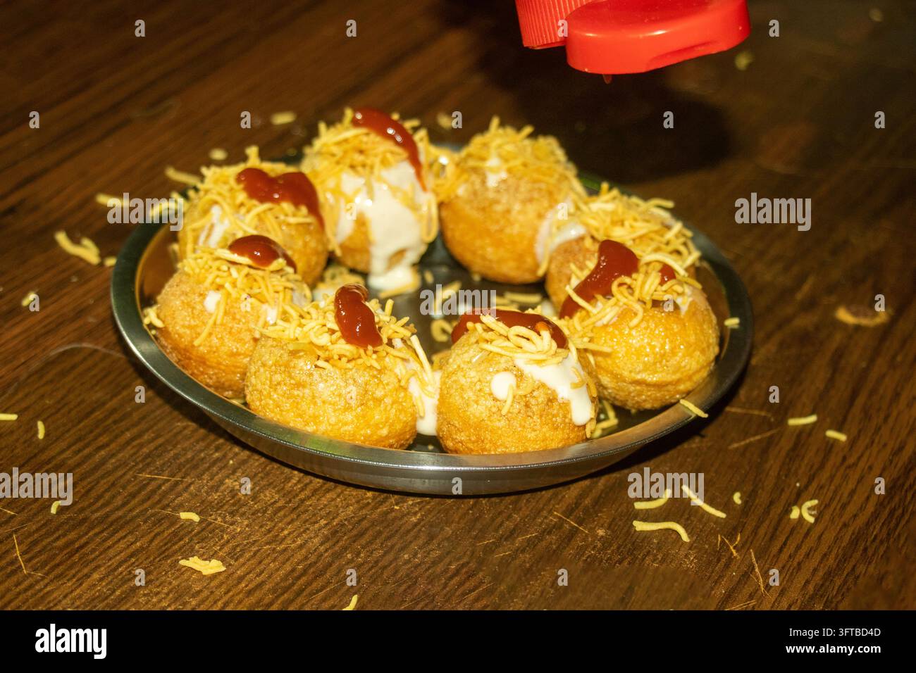 Teller mit knusprigen Semmelknödeln, belegt mit Joghurt, scharfer roter Chutney und gelben Sev-Nudeln, Snack serviert auf einem Holztisch im Restaurant. Stockfoto