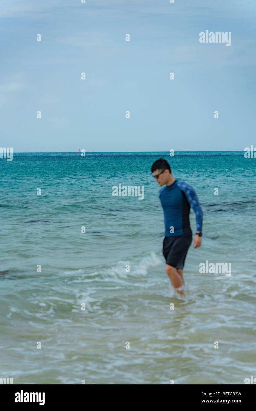 Mann in dunklem Rashguard und Shorts, der in ruhigem türkisfarbenem Meer watet, Con Dao, Vietnam Stockfoto