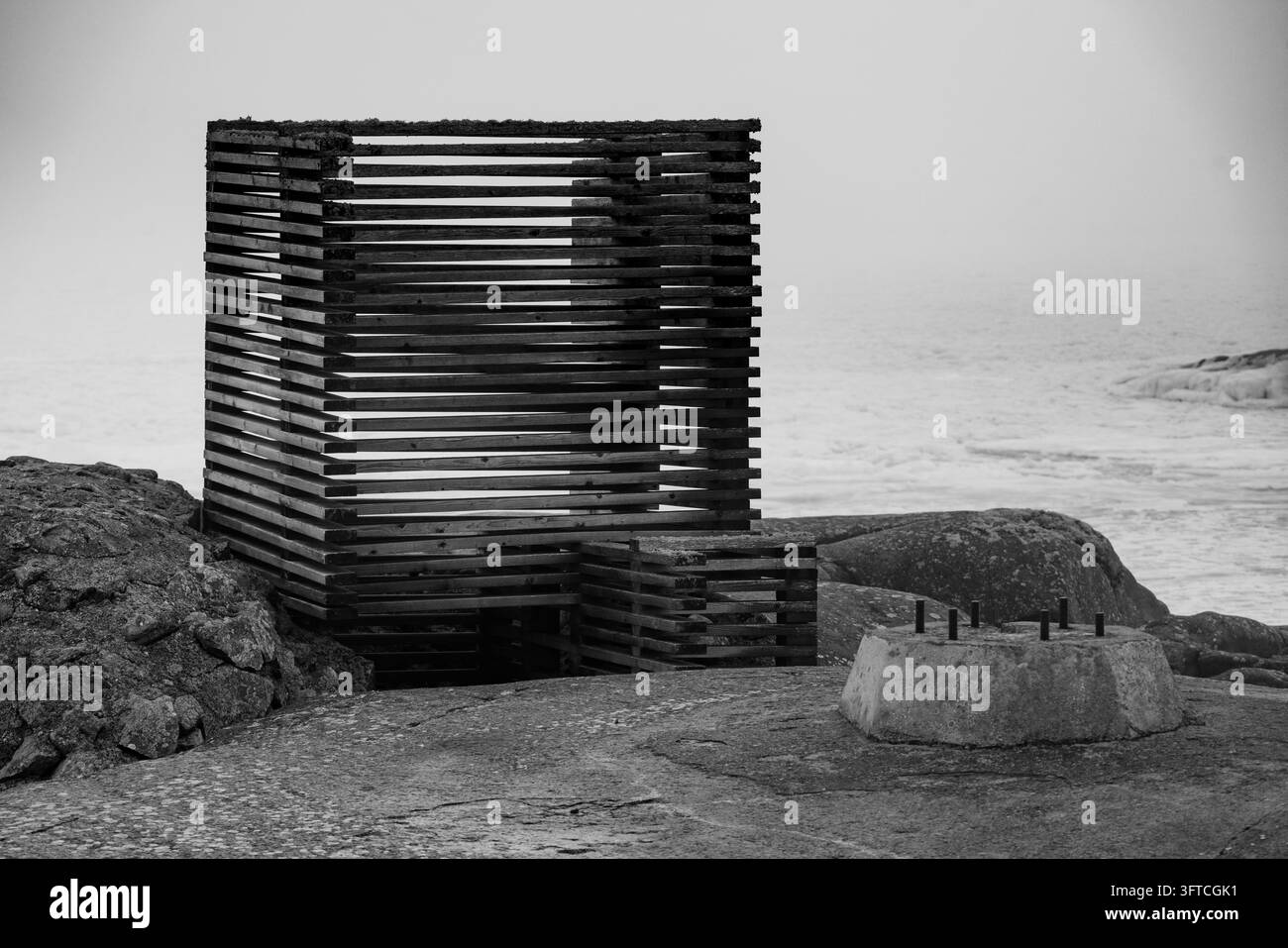 MONOCHROMANSICHT EINES KÜSTENKUNSTINSTALLATIONS, ENTFERNTE FINNISCHE INSEL IM TIEFEN WINTER, UTÖ, FINNLAND: Eine verwitterte, kistenartige Holz-Kunstinstallation und ein Betonblock ruhen auf einem felsigen Ufer, mit dem Meer im Hintergrund, in einer stimmungsvollen Monochrom-Darstellung. INFO: Utö ist eine entlegene finnische Insel im Schärenmeer südlich von Turku. Bekannt für seinen historischen Leuchtturm, raue Küstenlandschaft und maritimes Erbe, ist sie Finnlands südlichste bewohnte Insel und zieht Besucher an, die Ruhe, Vogelbeobachtung und spektakuläre Landschaften der Ostsee suchen. Stockfoto
