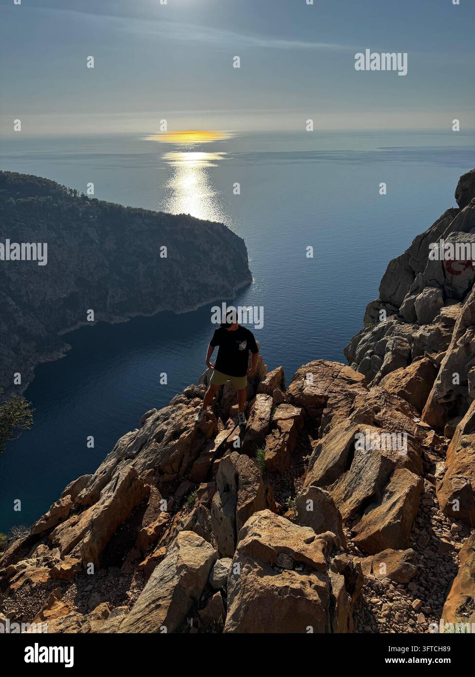 Wanderer auf einer felsigen Klippe über einer tiefblauen Bucht mit Sonnenfunkeln auf dem Meer, Lycische Weg nahe Faralya, Türkei - Smartphone-aufgenommenes Stockfoto