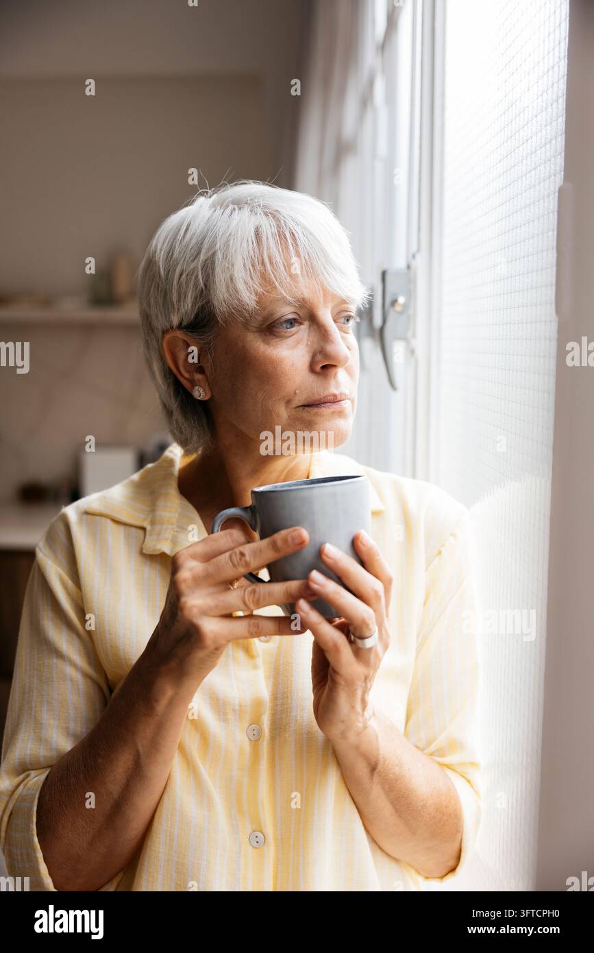 Seniorin schaut aus dem Fenster, hält eine Kaffeetasse, denkt und fühlt sich einsam Stockfoto