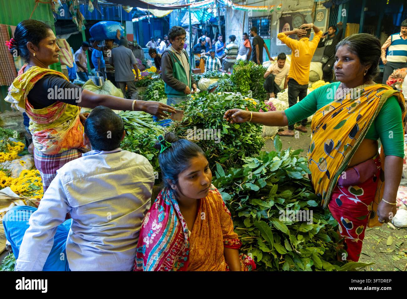 Verkäufer in gelben und roten Saris reichen Bündel mit Blattgemüse weiter, während Menschen auf dem Boden auf dem Blumenmarkt sitzen, West Bengal, Kalkutta, Indien Stockfoto