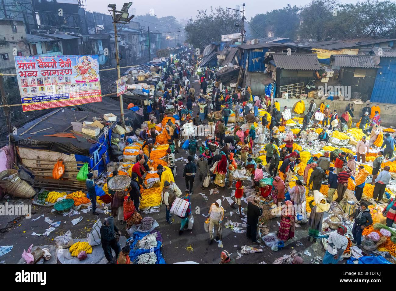 Hunderte von Käufern und Verkäufern füllen eine nasse Straße, die mit gelben und orangefarbenen Girlanden geschmückt ist, auf dem Blumenmarkt, West Bengal, Kalkutta, Indien Stockfoto