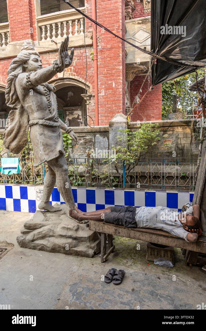 Mann liegt auf einer Holzbank neben einer großen Tonstatue einer stehenden Gottheit im Bezirk Kumartuli, West Bengal, Kalkutta, Indien Stockfoto