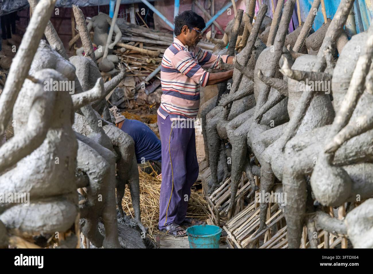 Mann in einem gestreiften Hemd steht zwischen unfertigen Tonskulpturen, während ein Arbeiter dahinter im Kumartuli-Bezirk, West Bengal, Kalkutta, Indien kniet Stockfoto
