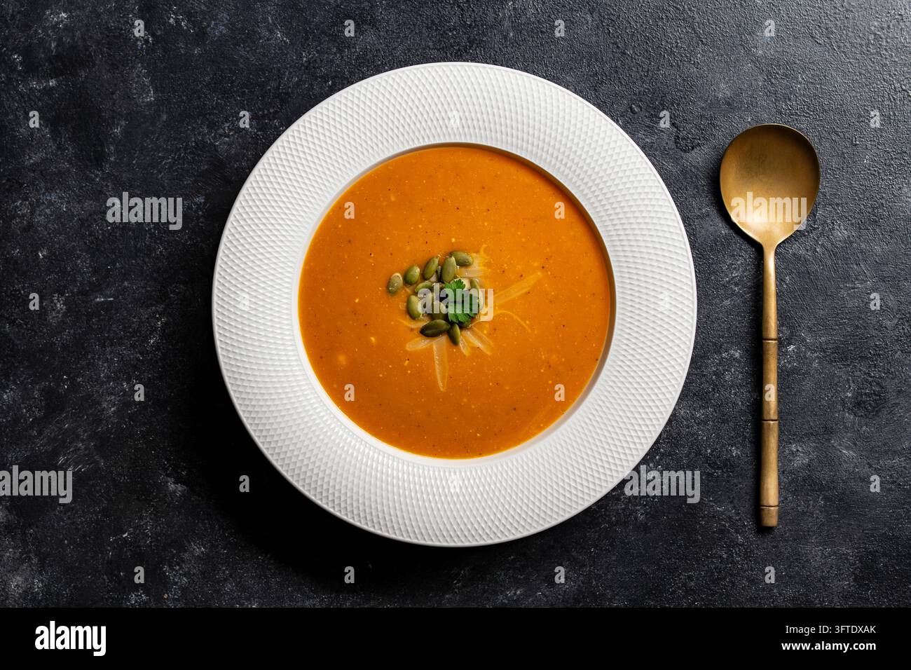 Kürbiskremesuppe auf dunklem Hintergrund mit goldenem Löffel. Oberansicht von orangefarbener Kürbis- oder Tomatenpüreesuppe in weißem Teller. Gesundes veganes Herbstkomfort Stockfoto