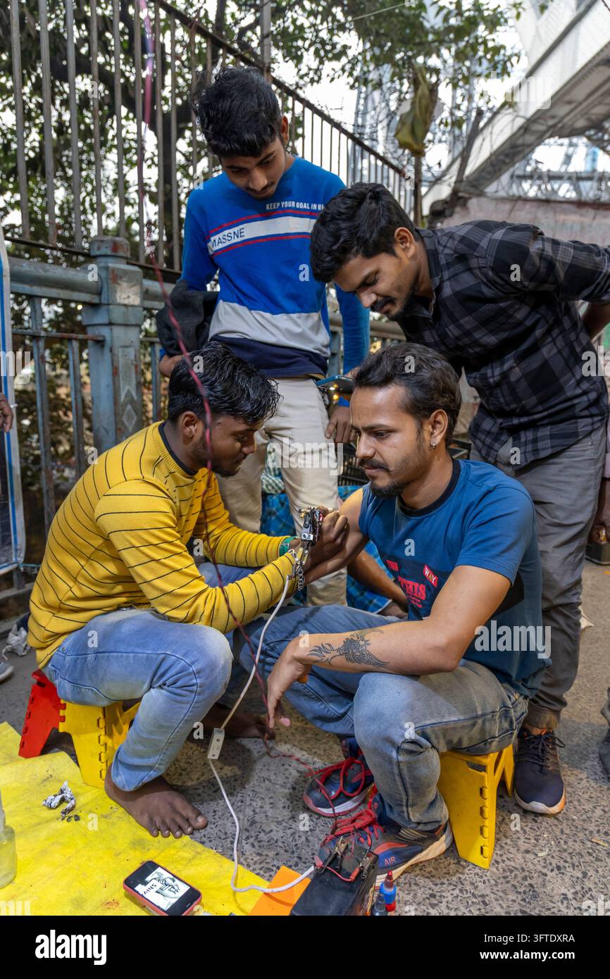 Ein Tätowierer arbeitet an der Howrah-Brücke, während drei andere sich neben einem Eisengeländer vorbeugen, um zuzusehen, West Bengal, Kalkutta, Indien Stockfoto