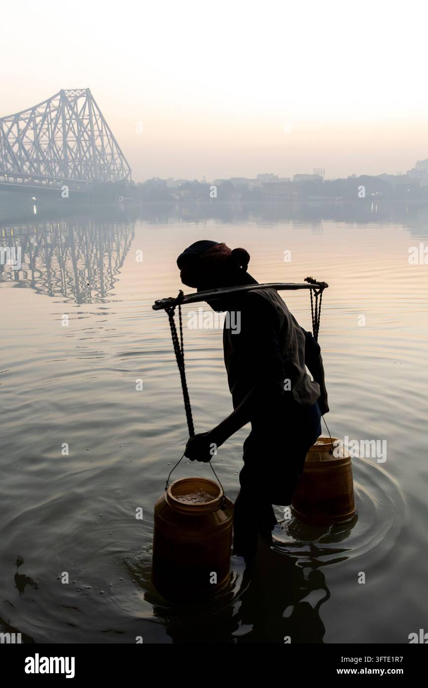 Wasserschaffner watet bei Morgengrauen in ruhiges Flusswasser und hält gelbe Krüge an einem Joch, während eine Stahlbrücke im Dunst verschwindet, Westbengalen, Kalkutta, Indien Stockfoto