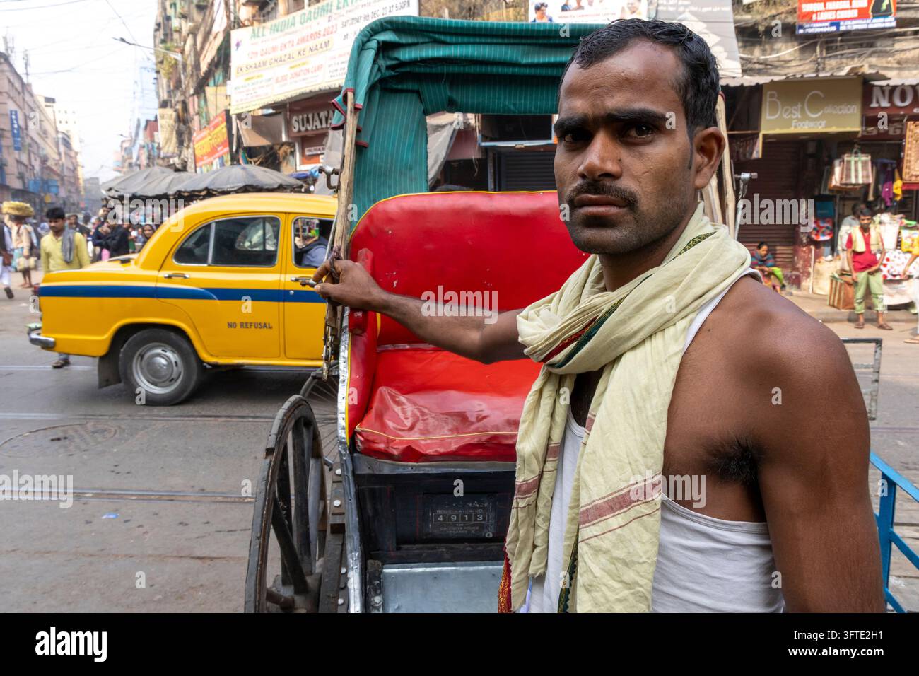 Ein ernster Mann mit einem cremefarbenen Schal und weißer Weste hält den Rahmen einer Rikscha, während gelbe Taxis die belebte Straße in Westbengalen, Kalkutta, Indien füllen Stockfoto