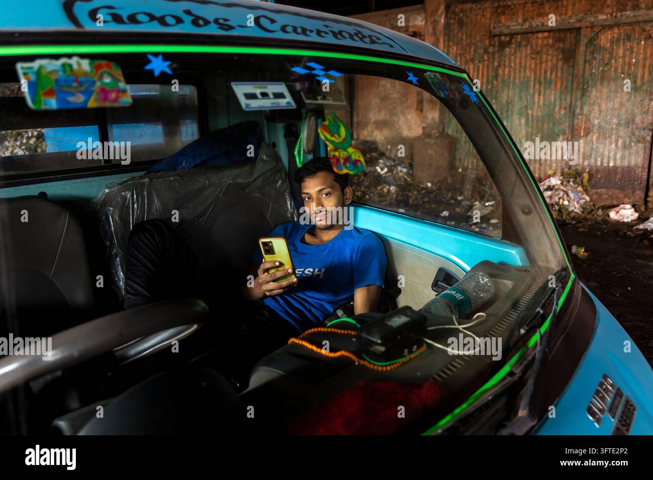Ein Fahrer lehnt sich in einem hellblauen Frachtfahrzeug zurück, das in einem heruntergekommenen Industriegebiet geparkt ist, und hält ein Smartphone, West Bengal, Kalkutta, Indien Stockfoto