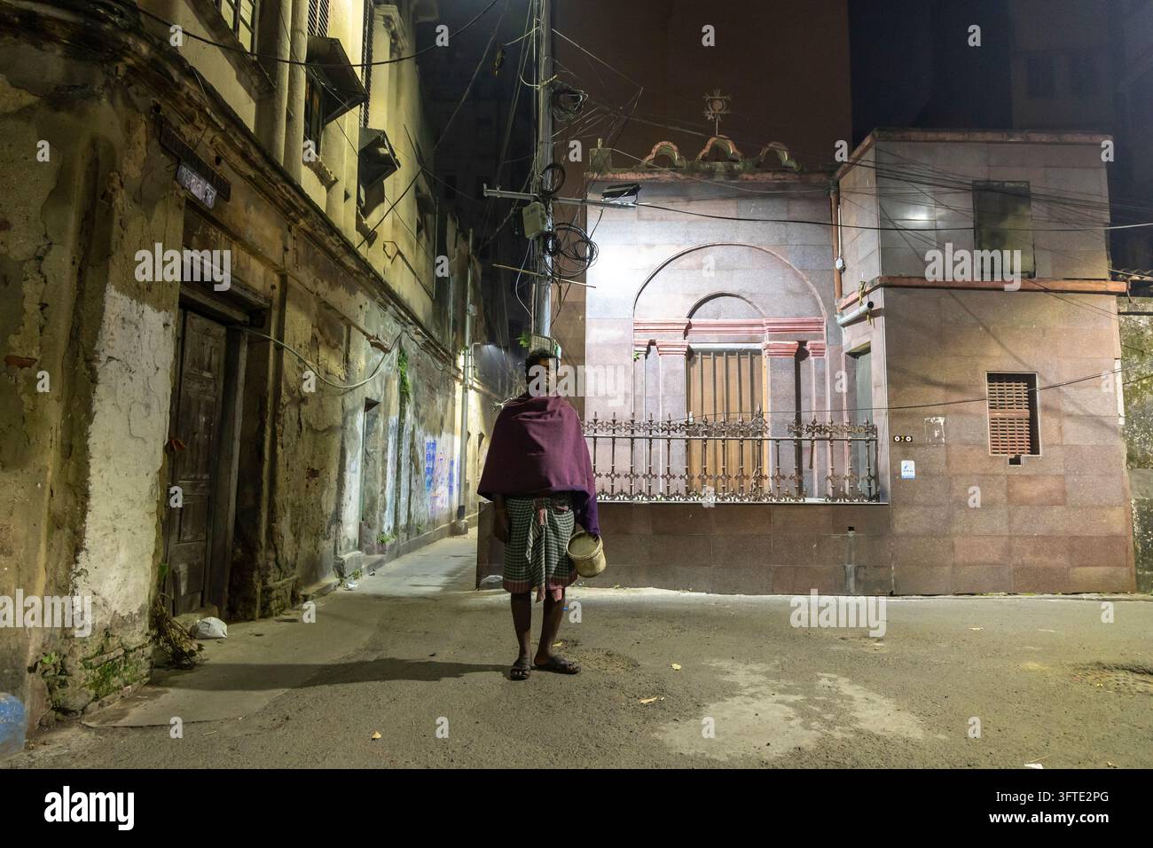 Eine kleine Figur, die einen Eimer hält, steht mitten auf einer leeren Gasse mit verwitterten Wänden und einer beleuchteten Struktur, West Bengal, Kalkutta, Indien Stockfoto