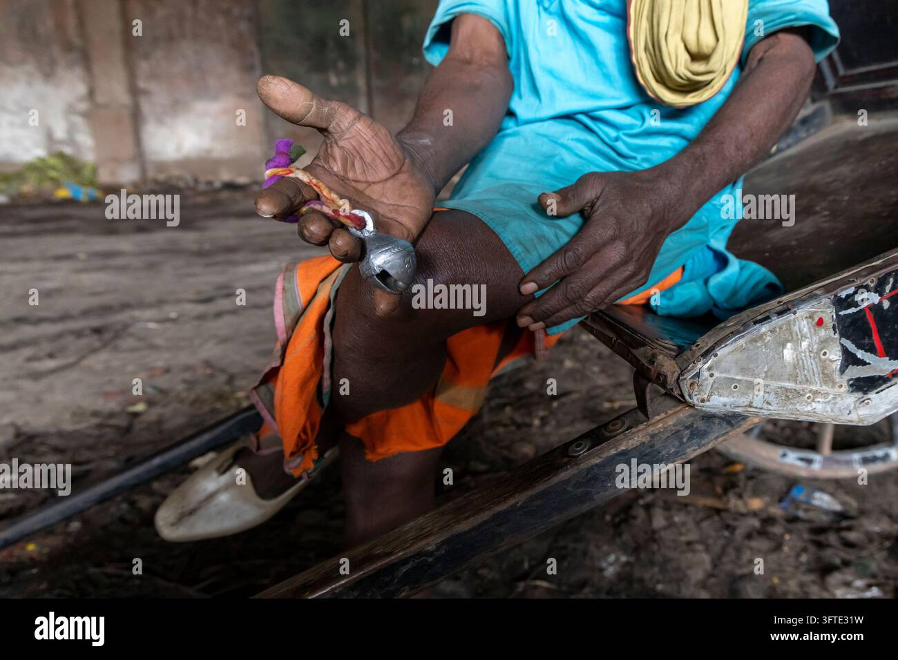 Nahaufnahme einer verwitterten Hand eines Mannes mit einer silbernen Glocke an einem lila und orangefarbenen Faden, die auf seinem Knie ruht, West Bengal, Kolkata, Indien Stockfoto