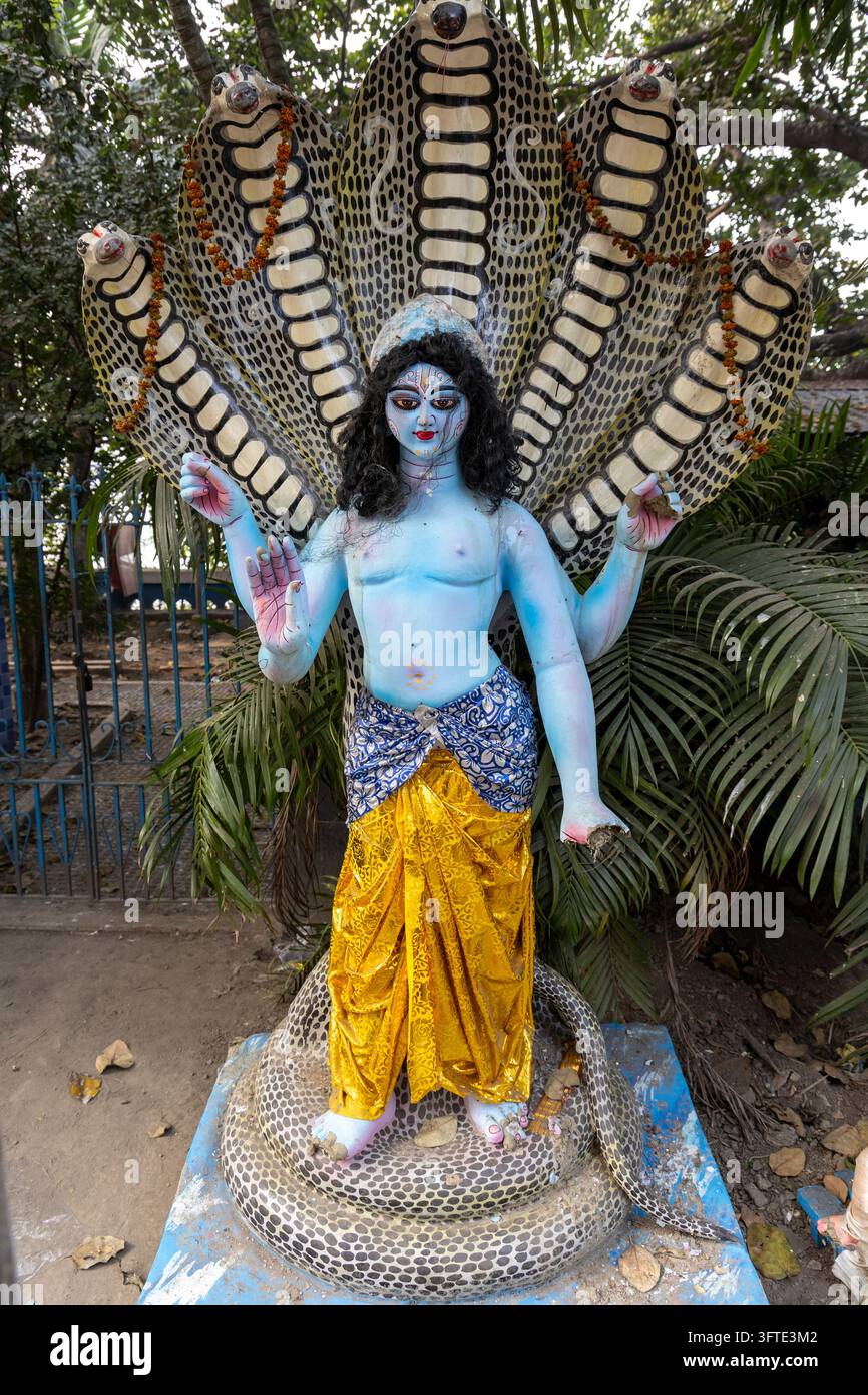 Straßenstatue einer Figur mit erhobenen Händen, die auf einer zusammengerollten Schlange vor einem Zaun und Palmenblättern steht, Westbengalen, Kalkutta, Indien Stockfoto