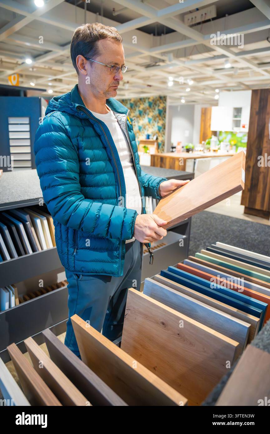 Ein Mann mit Brille betrachtet eine Holzmaserungsplatte neben einem Musterregal im Ausstellungsraum. Die Auswahl von Möbelbeschichtungen unterstützt die Planung von Heimrenovierungen. Stockfoto