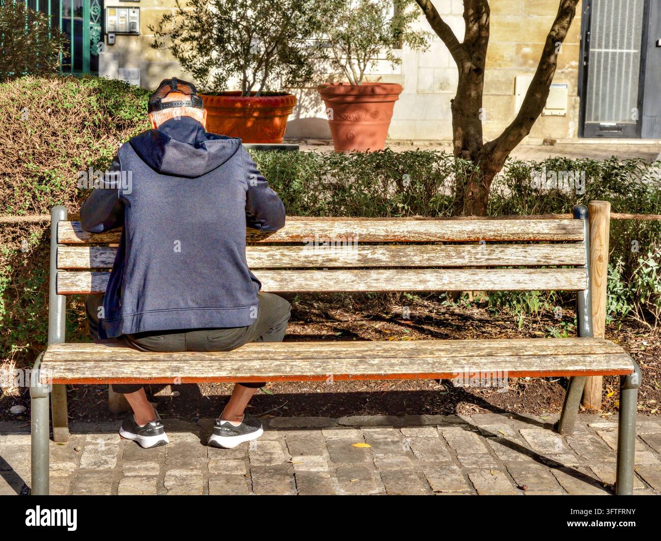 Älterer Mann in Kapuzenpullover und Baseballkappe, der mit dem Rücken nach vorne auf einer Bank sitzt und in einem öffentlichen Garten liest – Tours, Indre-et-Loire (37), Frankreich. Stockfoto