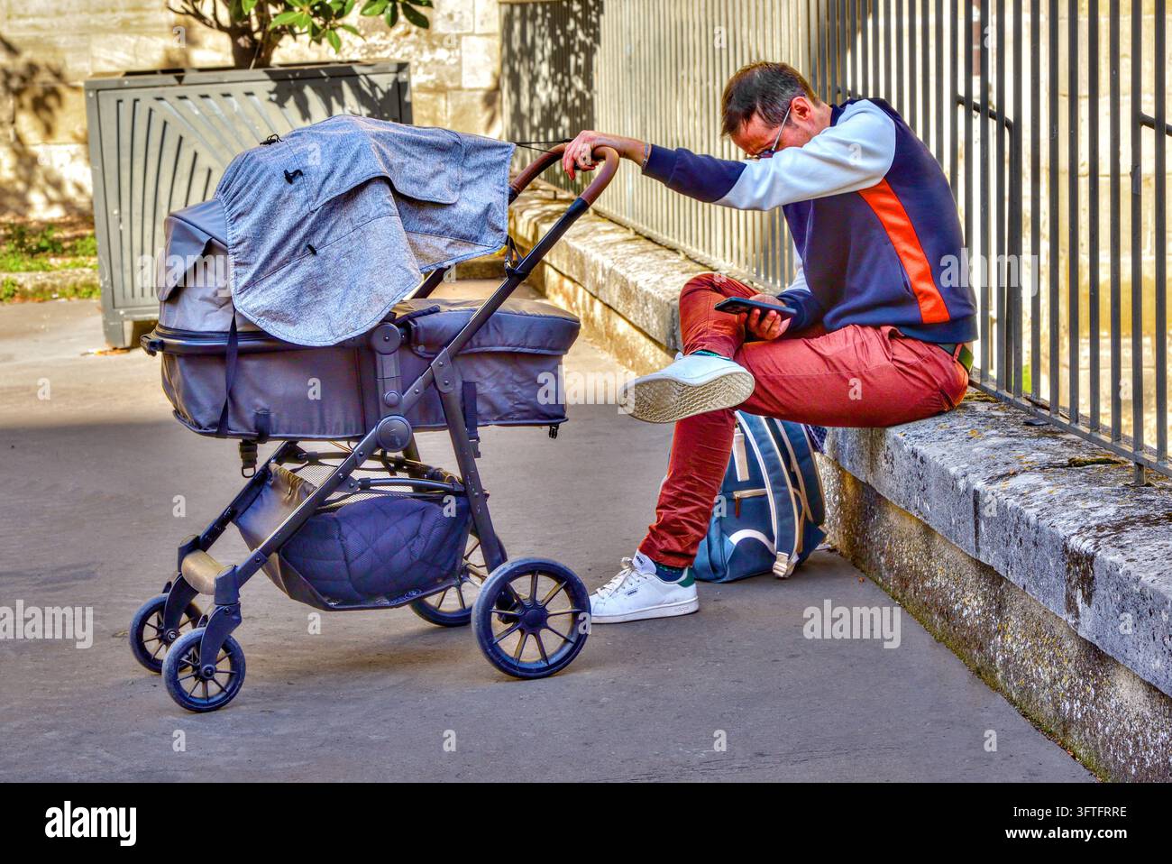 Mann sitzt auf niedriger Mauer und schaut auf Smartphone, während er ein Baby im Kinderwagen schaukelt – Tours, Indre-et-Loire (37), Frankreich. Stockfoto