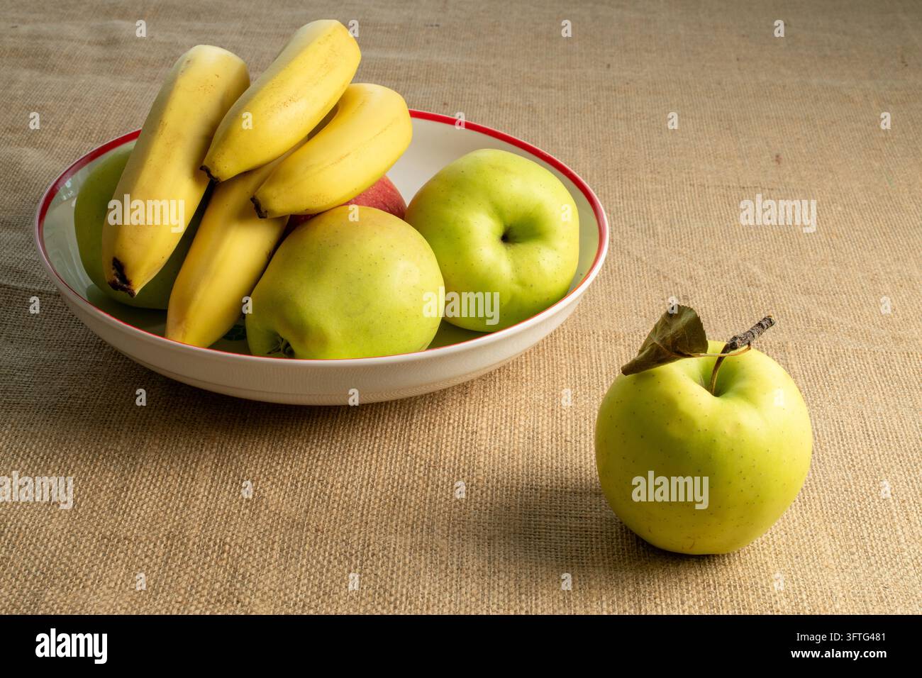 Frische gelbe Bananen und reife grüne und rote Äpfel in einer Keramikschale auf einem rustikalen Sackleinenhintergrund. Stockfoto